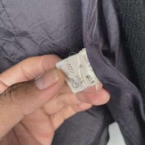 May include: Close-up of the interior lining of a dark gray jacket. A white tag is visible, with text and symbols. The lining is a smooth, dark gray fabric. The jacket's outer material is a textured, darker gray.