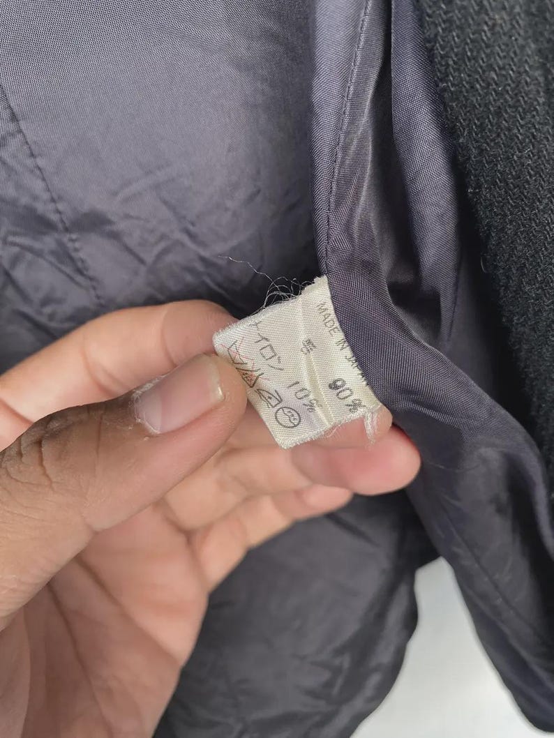 May include: Close-up of the interior lining of a dark gray jacket. A white tag is visible, with text and symbols. The lining is a smooth, dark gray fabric. The jacket's outer material is a textured, darker gray.