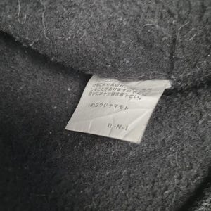 May include: Close-up of a dark gray fabric with a white tag attached. The tag has Japanese text and the code "&Pi;-&Nu;-1". The fabric appears to be a textured material, possibly wool or a similar textile.