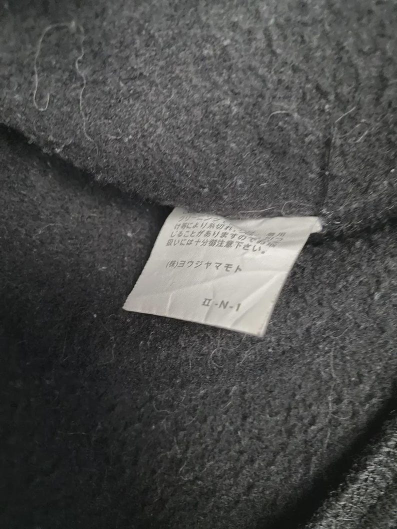 May include: Close-up of a dark gray fabric with a white tag attached. The tag has Japanese text and the code "&Pi;-&Nu;-1". The fabric appears to be a textured material, possibly wool or a similar textile.