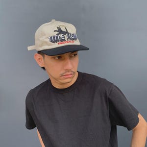 May include: A beige baseball cap with a black brim featuring an embroidered black cat graphic above the text "TITLE FIGHT" in white and "KINGSTON PA USA" in red. The cap has a structured crown and an adjustable strap closure at the back.