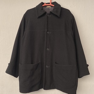 May include: A black wool coat with a classic design. The coat features a collared neckline, three buttons, two large front pockets, and long sleeves with folded cuffs. It hangs on a red hanger against a neutral background.
