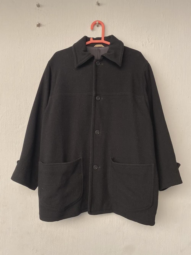 May include: A black wool coat with a classic design. The coat features a collared neckline, three buttons, two large front pockets, and long sleeves with folded cuffs. It hangs on a red hanger against a neutral background.