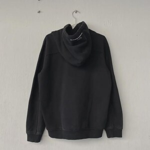May include: Black hoodie with a drawstring hood, long sleeves, and ribbed cuffs and hem. The back of the hoodie is plain, while the hood features white text. The hoodie is hanging on a metal hanger against a white wall.