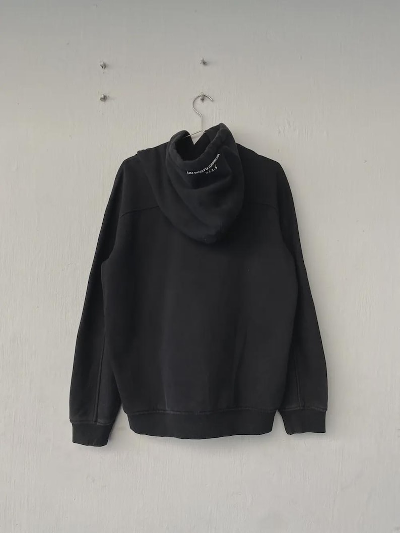 May include: Black hoodie with a drawstring hood, long sleeves, and ribbed cuffs and hem. The back of the hoodie is plain, while the hood features white text. The hoodie is hanging on a metal hanger against a white wall.
