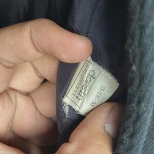 May include: Close-up of a tag inside a dark garment. The tag is rectangular with the text "dessin yohji yamamoto" and "D-TK-33" printed on it. The tag is white with black text.