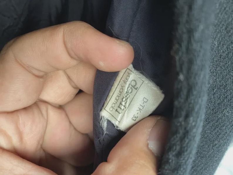 May include: Close-up of a tag inside a dark garment. The tag is rectangular with the text "dessin yohji yamamoto" and "D-TK-33" printed on it. The tag is white with black text.
