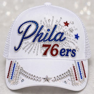 May include: White trucker hat with the word "Phila" in blue rhinestones and "76ers" in red and blue rhinestones. The hat features a mesh back, a curved brim, and decorative stars and rhinestones.