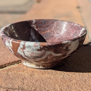 May include: A small, round bowl made of red, white, and brown marble. The bowl has a smooth, polished surface and is perfect for holding small items.