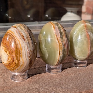 Op de afbeelding: Vier gepolijste onyx-eieren met verschillende patronen van bruine, groene en witte strepen. De eieren worden gepresenteerd op heldere standaards.
