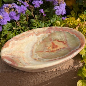 May include: An oval-shaped, natural stone dish with a polished finish. The dish features a blend of beige, cream, and reddish-brown hues, with a swirling pattern. The dish is surrounded by green and purple foliage.