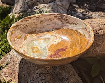 May include: A large, round, decorative bowl made of natural stone. The bowl has a swirling pattern of brown, white, and green colors. The bowl is sitting on a rock.