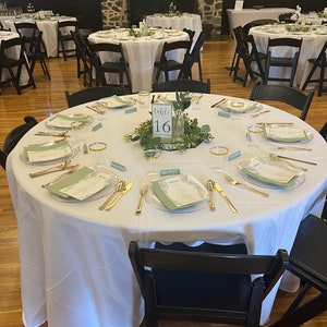 May include: A round white tablecloth with gold silverware, clear glasses, and green place cards. A centerpiece of greenery and a sign that reads "Table 16" is in the center of the table.