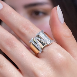 May include: A gold ring featuring a large, pave-set letter "M" design. The ring is worn on a person's finger.