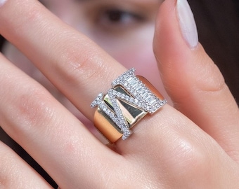 Può includere: Un anello d'oro con un grande design a forma di lettera "M" incastonato a pavé. L'anello è indossato su un dito.