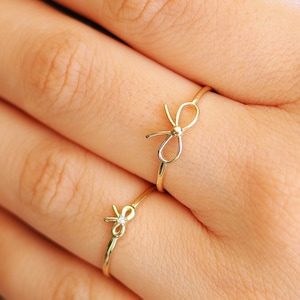 Op de afbeelding: Twee gouden ringen met een strikdesign. De ringen worden gedragen aan de wijs- en middelvinger van een hand.