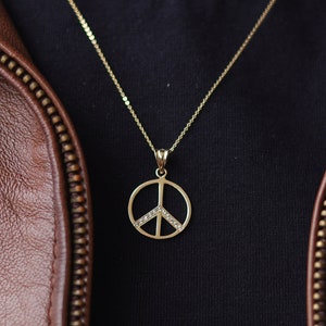 May include: A gold peace sign pendant necklace with small clear stones on a gold chain. The necklace is worn over a black t-shirt and a brown leather jacket.