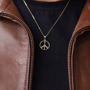 May include: A gold peace sign pendant necklace with small clear stones on a chain. The necklace is worn over a black t-shirt and brown leather jacket.