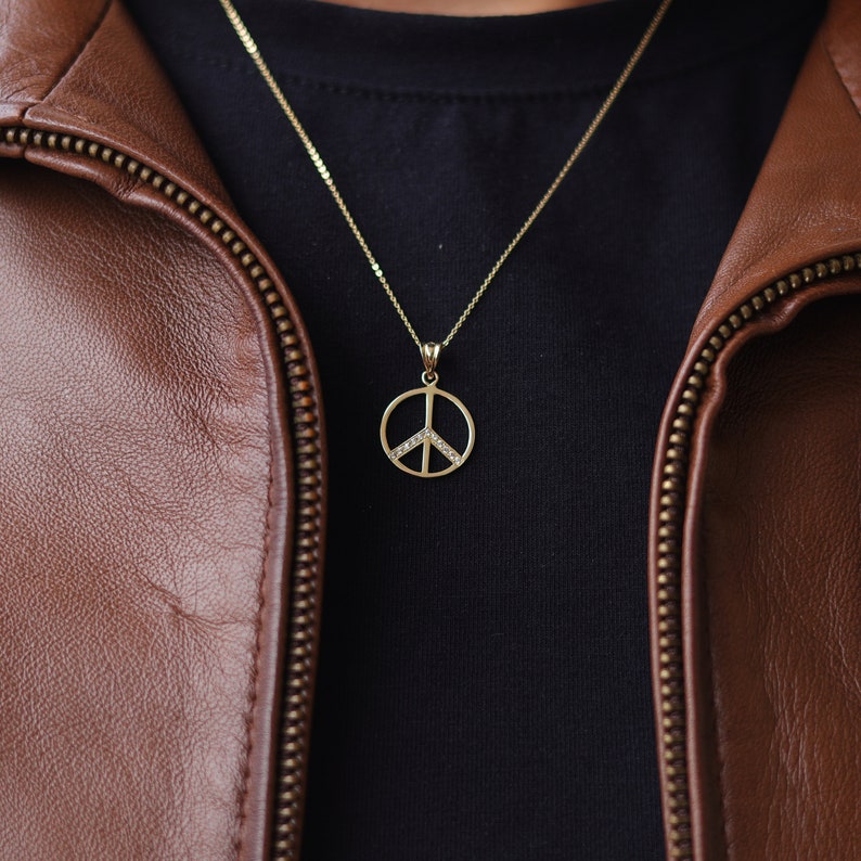 May include: A gold peace sign pendant necklace with small clear stones on a chain. The necklace is worn over a black t-shirt and brown leather jacket.