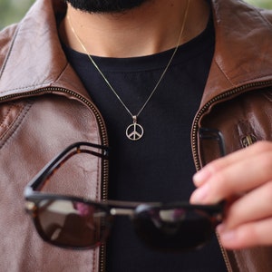 May include: A gold chain necklace with a peace sign pendant. The necklace is worn by a person wearing a brown leather jacket and a black t-shirt.