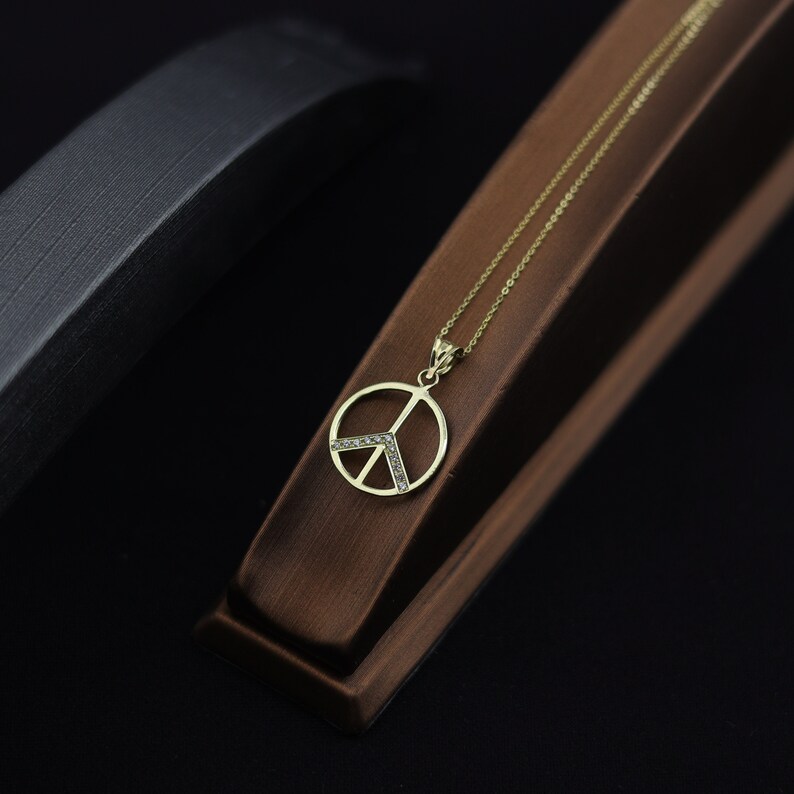 May include: A gold peace sign pendant necklace with small clear stones set around the edges. The necklace is displayed on a brown wooden stand.