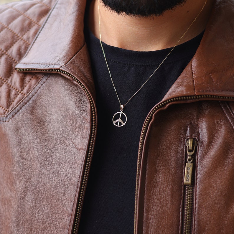 May include: A gold peace sign pendant necklace on a gold chain. The necklace is worn by a person wearing a brown leather jacket.