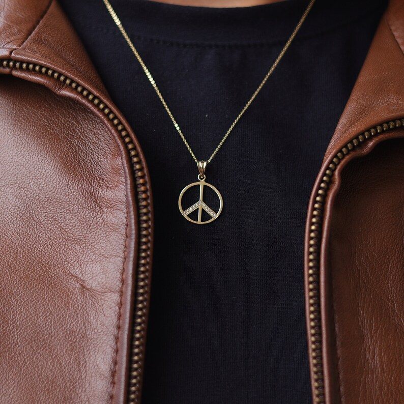 May include: A gold peace sign pendant necklace with small clear stones on a gold chain. The necklace is worn over a black t-shirt and a brown leather jacket.