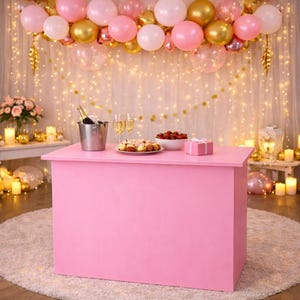 May include: A rectangular pink event table featuring a silver champagne bucket, two glass flutes, a plate of assorted pastries, a bowl of strawberries, and a small pink gift box. The table is set against a backdrop of pink and gold balloons.