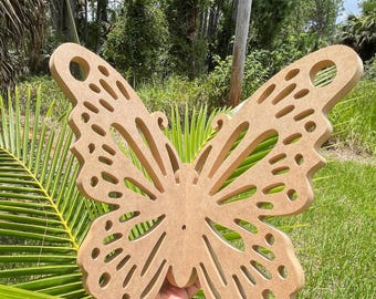 May include: A large, intricately cut wooden butterfly with detailed wing patterns. The butterfly is a light brown color and has a smooth finish. The image is taken outdoors with green foliage in the background.