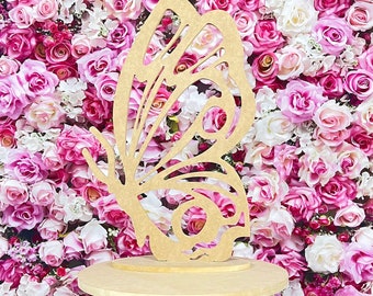May include: A wooden butterfly cutout with intricate details, standing on a wooden pedestal against a backdrop of pink and white artificial roses.