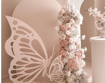 May include: Two large, white, cutout butterfly shapes stand on a light pink floor. The larger butterfly is in the foreground and the smaller butterfly is behind it. The butterflies are in front of a light pink wall with a large, light pink circle behind the larger butterfly. A white pillar with a white floral arrangement is to the right of the butterflies.