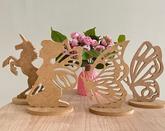 May include: Four wooden cutout decorations: a unicorn, a fairy, and two butterflies. The unicorn and fairy are brown, and the butterflies are a lighter brown. The decorations are on a wooden table with a pink vase of flowers in the background.