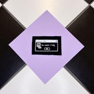 Op de afbeelding: Een zwart patch met witte tekst en een computergrafiek, met de tekst "Hardware conflict" en "Your computer is lonely." De patch ligt op een lichtpaarse vierkant, tegen een zwart-wit geblokte achtergrond.