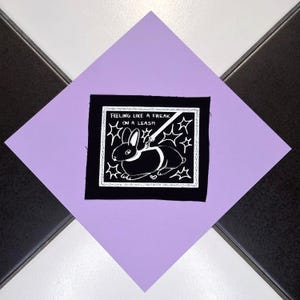 Könnte beinhalten: Ein schwarz-weißer Aufnäher mit einem Hasen an der Leine und dem Text "Feeling like a freak on a leash". Der Aufnäher befindet sich auf einer lavendelfarbenen Raute, vor einem schwarz-weiß gefliesten Hintergrund.