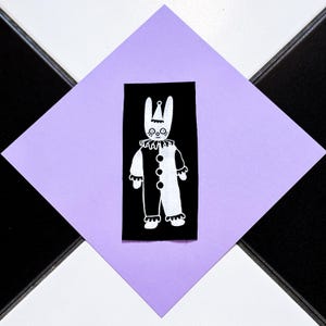 Op de afbeelding: Zwart-wit illustratie van een konijn verkleed als clown, met een gerimpelde kraag en een pak met knopen. De afbeelding staat op een zwarte rechthoek, geplaatst op een paarse ruit, met een zwart-witte dambord achtergrond.