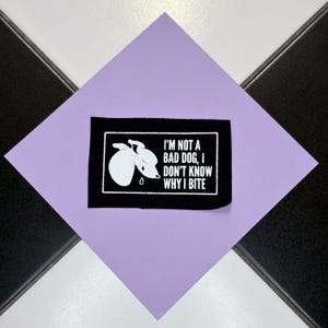 Peut inclure: Un patch noir avec du texte blanc et une illustration de chien. Le texte se lit comme suit : « I'M NOT A BAD DOG, I DON'T KNOW WHY I BITE ». Le patch est posé sur un fond violet clair, avec un motif à carreaux noir et blanc en arrière-plan.