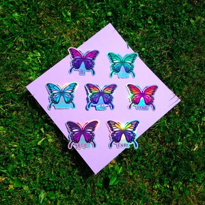 Può includere: Sette adesivi a forma di farfalla iridescenti con vari colori, tra cui viola, blu e arancione, sono disposti su un quadrato lavanda. Ogni farfalla ha una parola stampata sotto: "Bi", "Fag", "Dyke", "Ace" e "Enby". Gli adesivi sono su erba verde.
