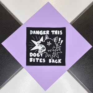 Peut inclure: Un écusson carré noir avec un texte blanc et un dessin de chien qui grogne. Le texte dit "DANGER THIS DOG BITES BACK" avec des points d'exclamation. L'écusson est sur un carré lavande, sur un fond à carreaux noir et blanc.