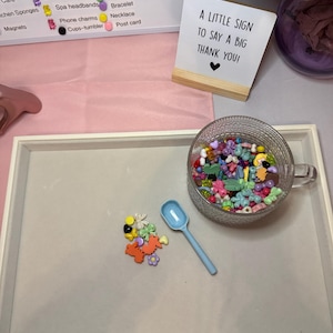 May include: A clear glass cup filled with colorful beads and charms, alongside a light blue scoop and a small pile of charms. A sign reads "A LITTLE SIGN TO SAY A BIG THANK YOU!" on a wooden base. A list of craft supplies is in the background.