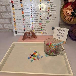 Peut inclure: Un plateau blanc contient une tasse en verre transparente remplie de bibelots colorés et une petite cuillère bleue. Un tableau blanc répertorie divers articles tels que du brillant à lèvres, des autocollants et des colliers. Une petite pancarte indique "A LITTLE SIGN TO SAY A BIG THANK YOU!"