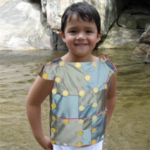 May include: A patchwork vest in shades of blue, gray, and green, adorned with gold-colored circular accents. The vest has a square neckline and short sleeves, with red trim. The garment is worn by a child.