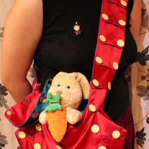 May include: A red satin tote bag with large yellow polka dots and a matching strap. The bag holds a stuffed bunny clutching an orange carrot. The strap is adorned with yellow circular accents.