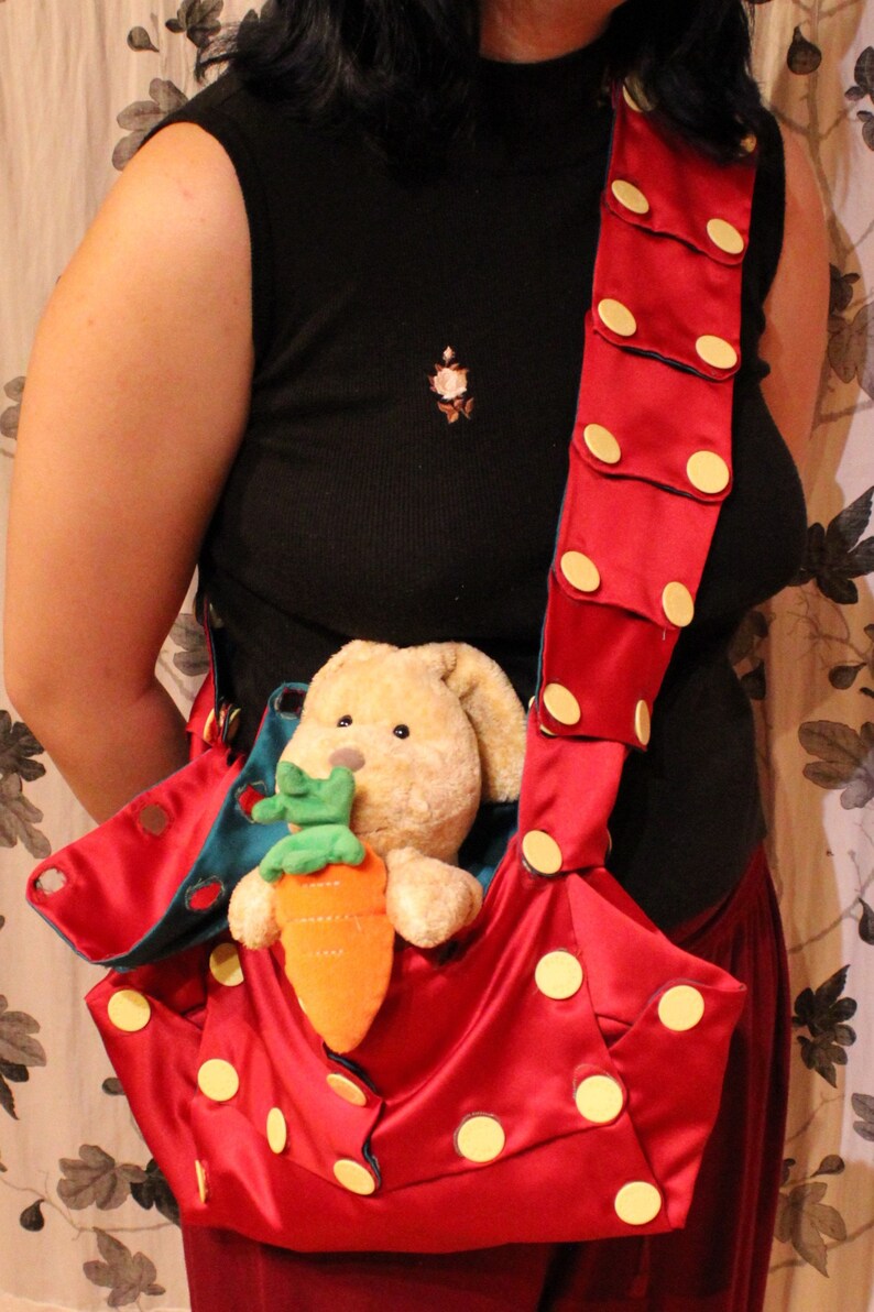 May include: A red satin tote bag with large yellow polka dots and a matching strap. The bag holds a stuffed bunny clutching an orange carrot. The strap is adorned with yellow circular accents.