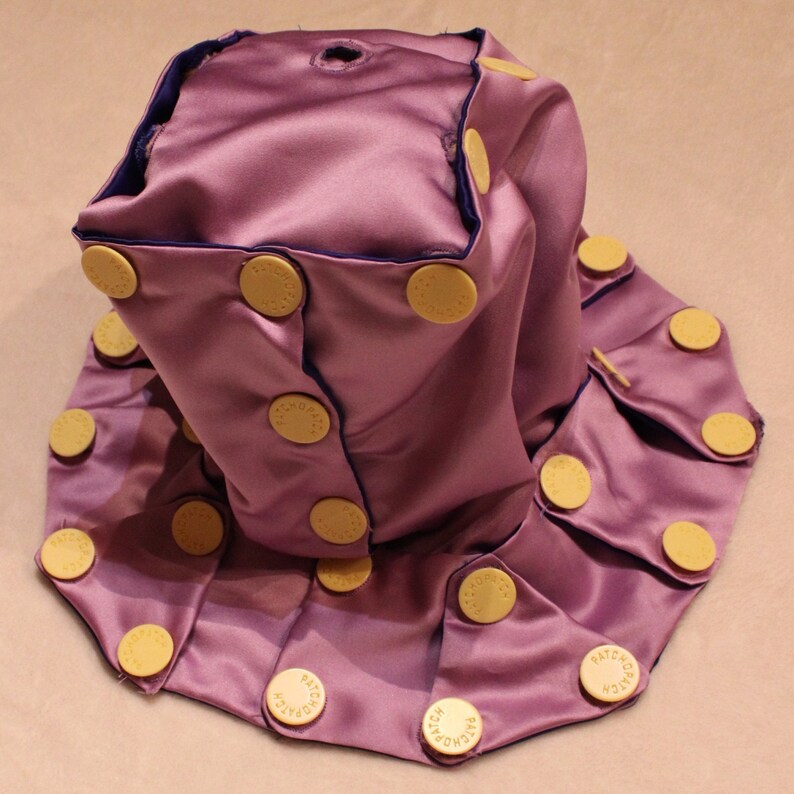 May include: A lavender satin hat with a dark blue trim and multiple yellow buttons. The buttons are circular and have the word "PATCH" printed on them. The hat has a unique, folded design.