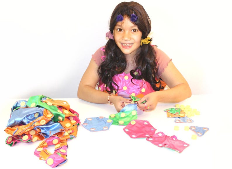 May include: A young person is seated at a white table, assembling colorful fabric shapes. The shapes are in various colors, including blue, green, orange, and pink, with yellow polka dots. The person is wearing a pink top with yellow polka dots.