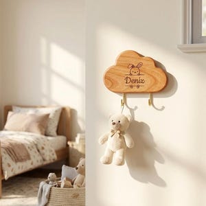 Puede incluir: Un perchero de pared de madera con forma de nube con el nombre "Deniz" grabado debajo de una ilustración de un conejito. Dos ganchos de latón cuelgan debajo. Un pequeño osito de peluche beige con un lazo está suspendido de un gancho.