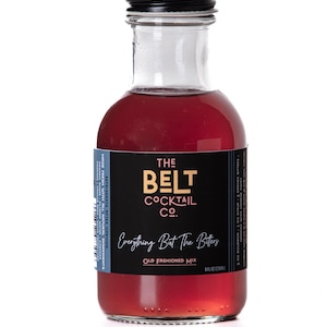 May include: A glass bottle of The Belt Cocktail Co. Everything But The Bitters Old Fashioned Mix. The bottle is filled with a red liquid and has a black label with white text.