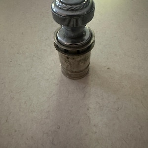 May include: A vintage car cigarette lighter with a silver finish. The top has a decorative design, and the body is cylindrical. The lighter is made of metal and shows signs of age, suggesting it is a classic car accessory.