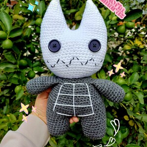 K&ouml;nnte beinhalten: Ein geh&auml;keltes Pl&uuml;schtier in Form einer Katze mit wei&szlig;em Kopf und grauem K&ouml;rper. Das Spielzeug hat gro&szlig;e schwarze Knopfaugen und einen gestickten Mund. Das Spielzeug wird in einer Hand vor einem gr&uuml;nen, belaubten Hintergrund gehalten.