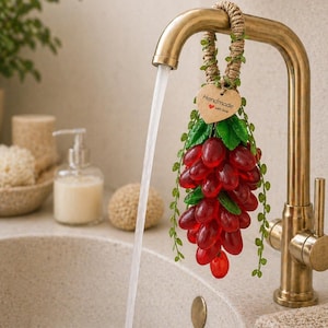 May include: A cluster of red, translucent grape-shaped soap hanging from a brass tap by a jute rope. A heart-shaped tag attached to the rope features the text "Handmade with love" and a small red heart icon. Green plastic leaves accent the grapes.
